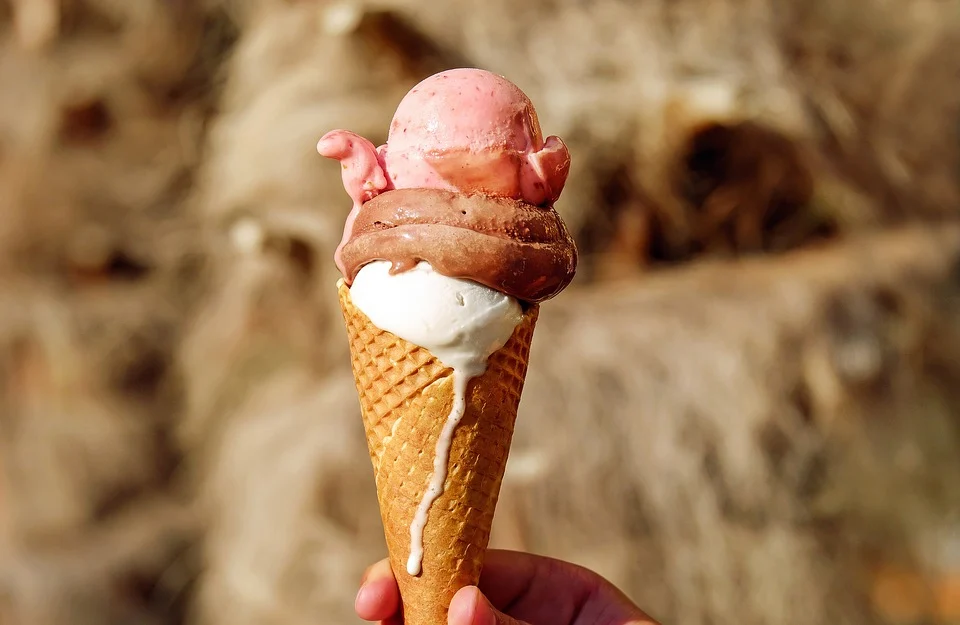 an ice cream cone with three scoops of ice cream is held by a hand in the middle of the picture. The ice cream scoops start to melt in the sun.