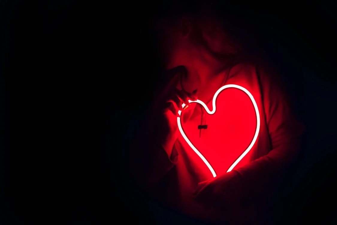 a woman stands in a dark room carrying a heart made of a red neon tube