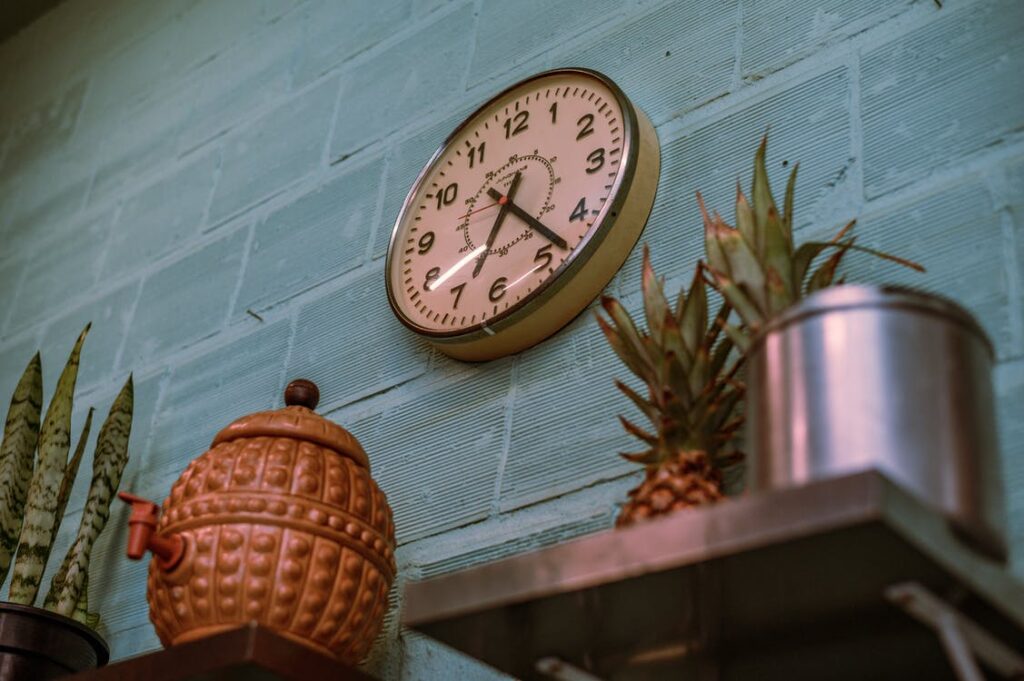 Eine Uhr hängt an einer blauen Wand, darunter ist ein Regalbrett mit einer Kanne und einer Ananas