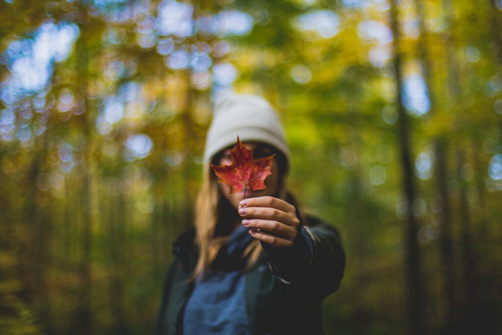 Junge Frau mit grauer Mütze hält verwelktes Herbstblatt in die Kamera vor ihr Gesicht