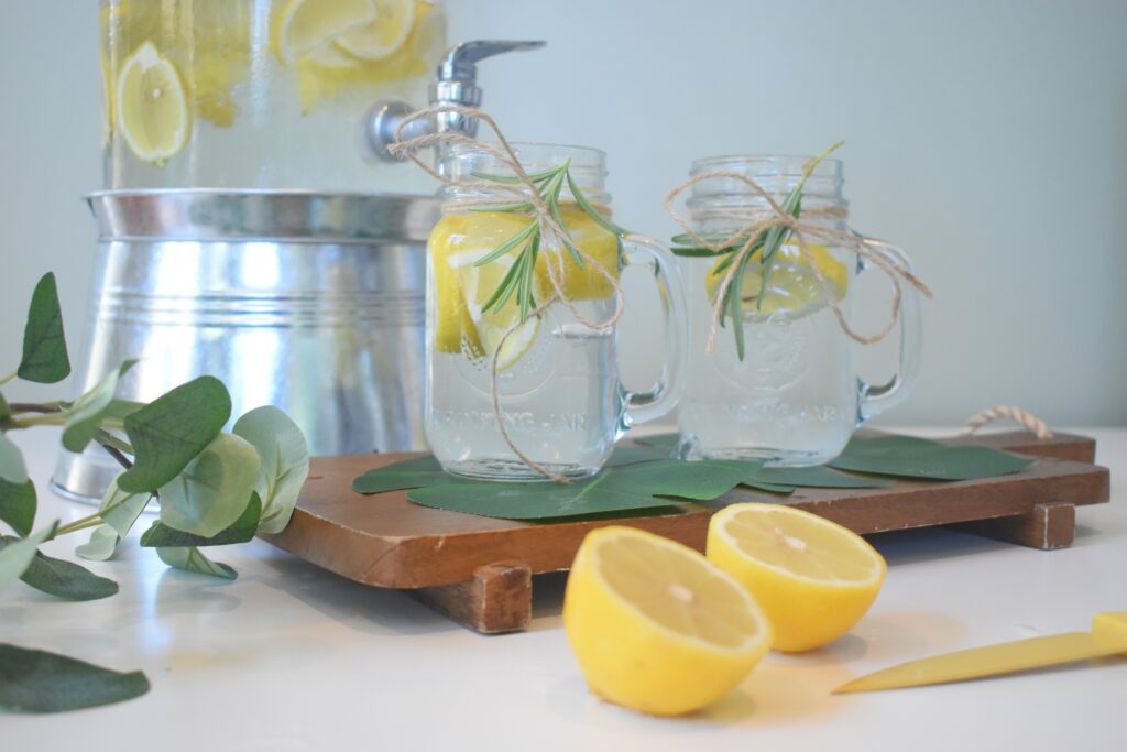 Ein Glasfass mit Zitronenwasser, in dem Zitronenscheiben schwimmen, davor stehen zwei Gläser mit Henkel, die mit Zitronenwasser und Zitronenscheiben gefüllt sind, auf einem Holzpodest, im Vordergrund liegen zwei halbe Zitronen