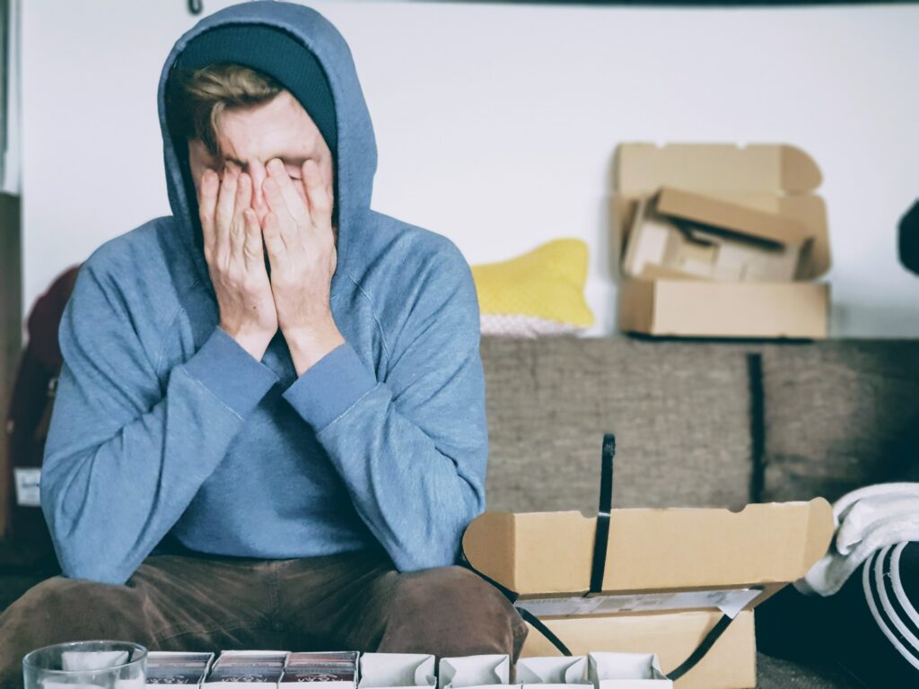 Boy in blue hoodie holds his hands in front of his face, in the background is cardboard packaging material