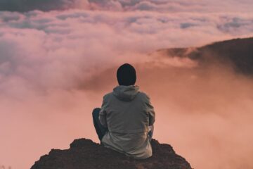 Junge mit schwarzer Mütze sitzt auf einem Fels über den Wolken und genießt die Aussicht
