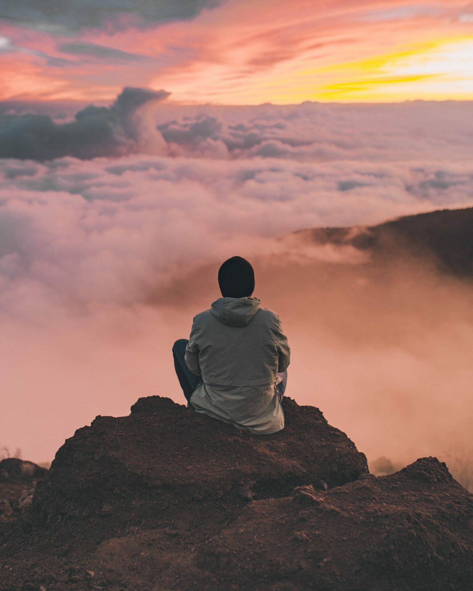 Boy with black cap sits on a rock above the clouds and enjoys the view