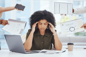 Woman sits stressed at her desk with her hands on her head. She is given many different tasks from outside.