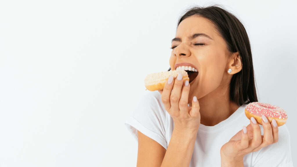 Brunette woman bites with pleasure in donut with beige glaze and sprinkles