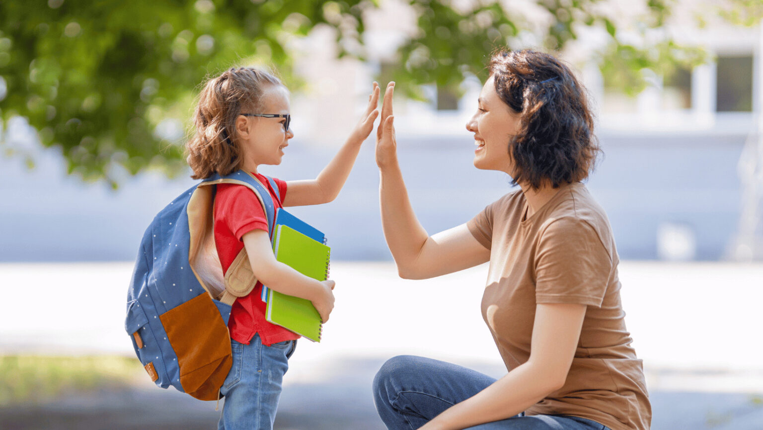 Brünette Mutter und ein kleines Schulkind mit Brille, Schulbüchern und blauem Rucksack geben sich lächelnd ein Highfive