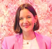 Brunette woman in a pink blazer and white tshirt smiles at the camera. In the background is a wall of roses.