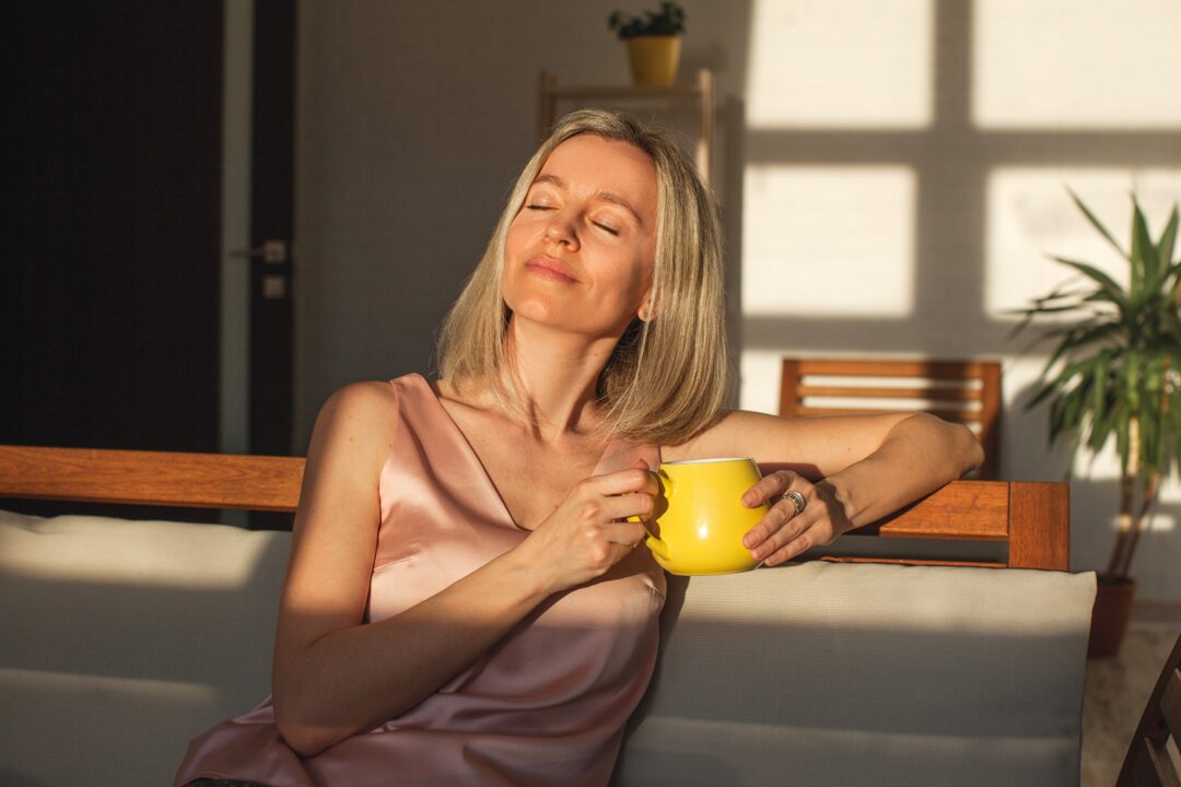 Woman sitting relaxed on sofa with sunshine on her face. She has a cup in her hand
